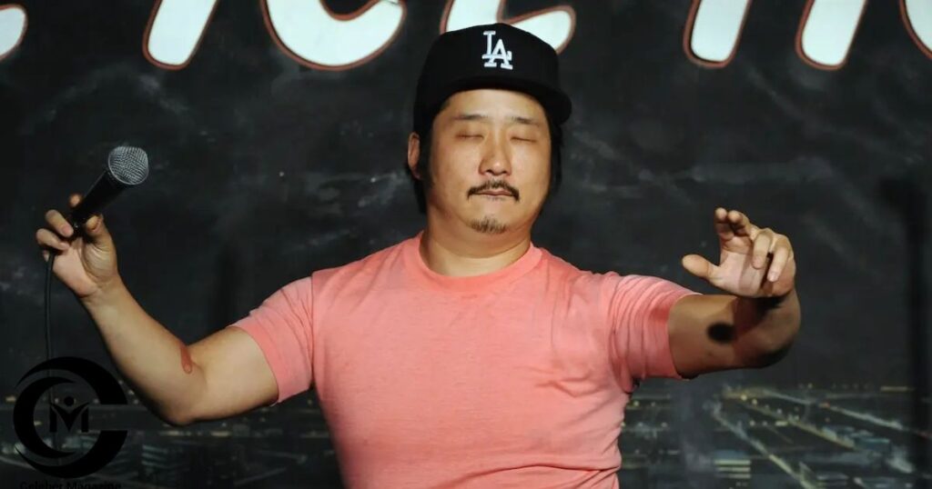 Bobby Lee performing stand-up comedy on stage wearing a pink shirt and black LA cap with a microphone in hand.