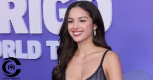 Olivia Rodrigo smiling at a red carpet event in a dark dress, standing in front of a purple backdrop.